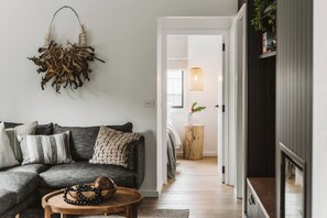 A modern living room with a dark grey sofa, a round wooden coffee table, and a woven wall hanging. A wooden stool with a plant is visible in the background.