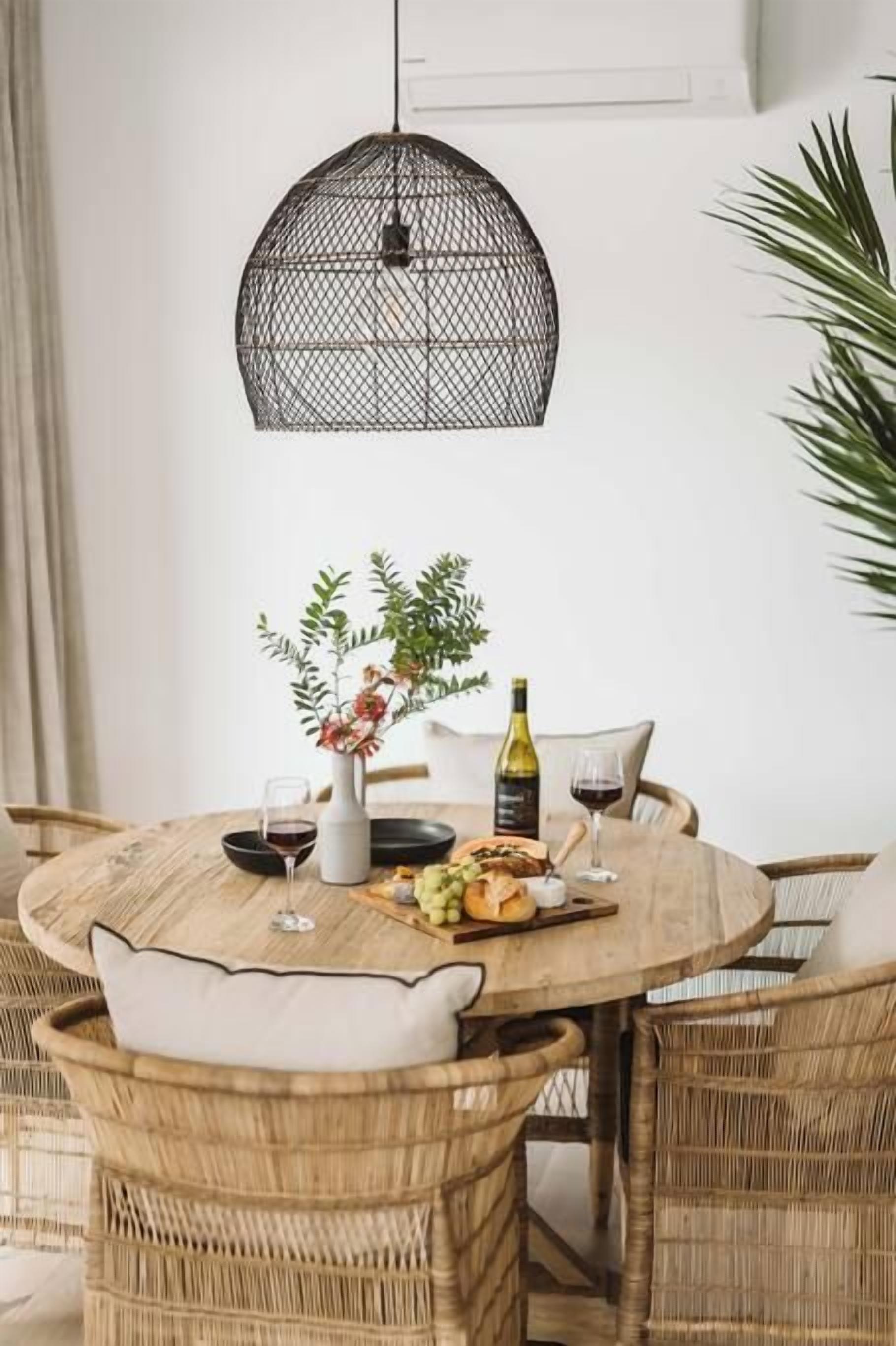 A round wooden dining table with wicker chairs, a black metal pendant light, and a small potted plant.