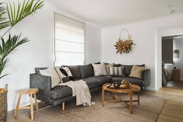 A modern living room with a grey sectional sofa, a round wooden coffee table, a small wooden stool, a large potted plant, and a hanging decorative piece. - The Lull Culburra (Culburra Beach)