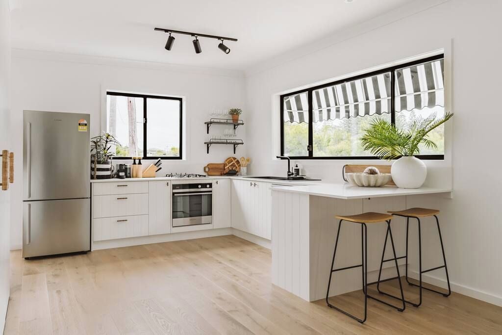 Een moderne keuken met witte kasten, een inbouwoven, een koelkast en een raam met uitzicht. Op het aanrecht staan twee barkrukken en een potplant.