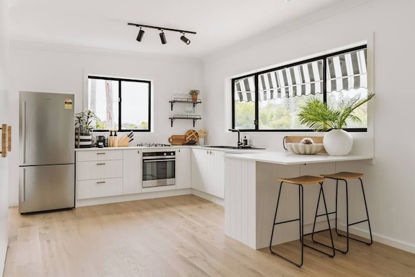 A modern kitchen with white cabinetry, a built-in oven, a refrigerator, and a window with a view. There are two stools and a potted plant on the counter.