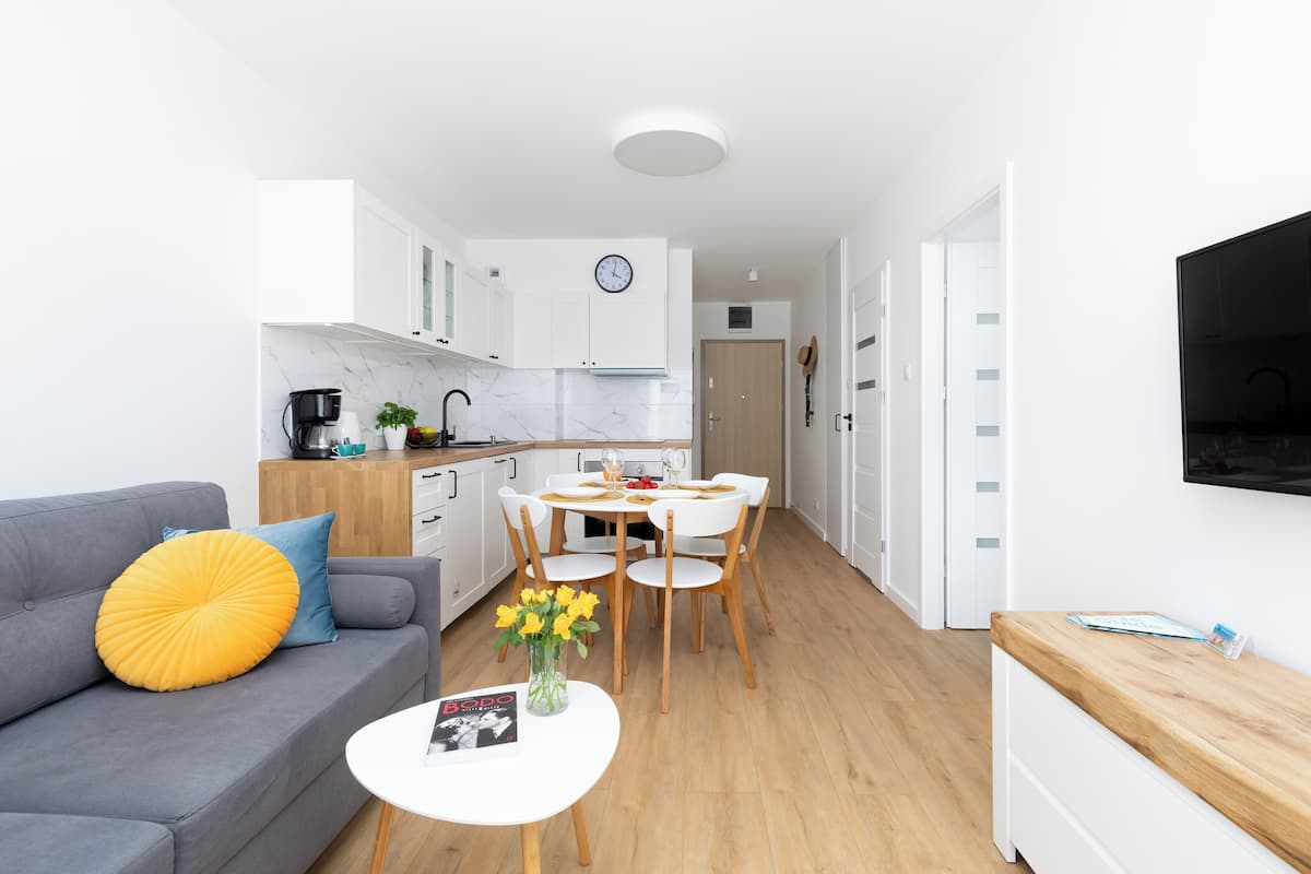A modern living room with a grey sofa, a small white table with a magazine, and a yellow pillow. A dining area with a white table and chairs is visible in the background.