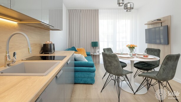 Uma cozinha moderna com pia embutida, sala de jantar com mesa redonda e cadeiras, e área de estar com sofá azul e televisão fixada na parede.