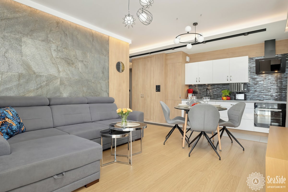 A modern living room with a grey sofa, a small round table with a vase of flowers, and a dining area with grey chairs and a white table.