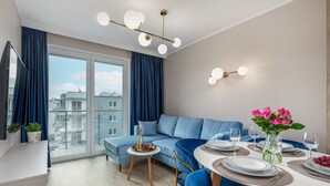 Uma sala de estar moderna com um sofá azul, uma mesa de jantar posta para dois e uma grande janela com cortinas.