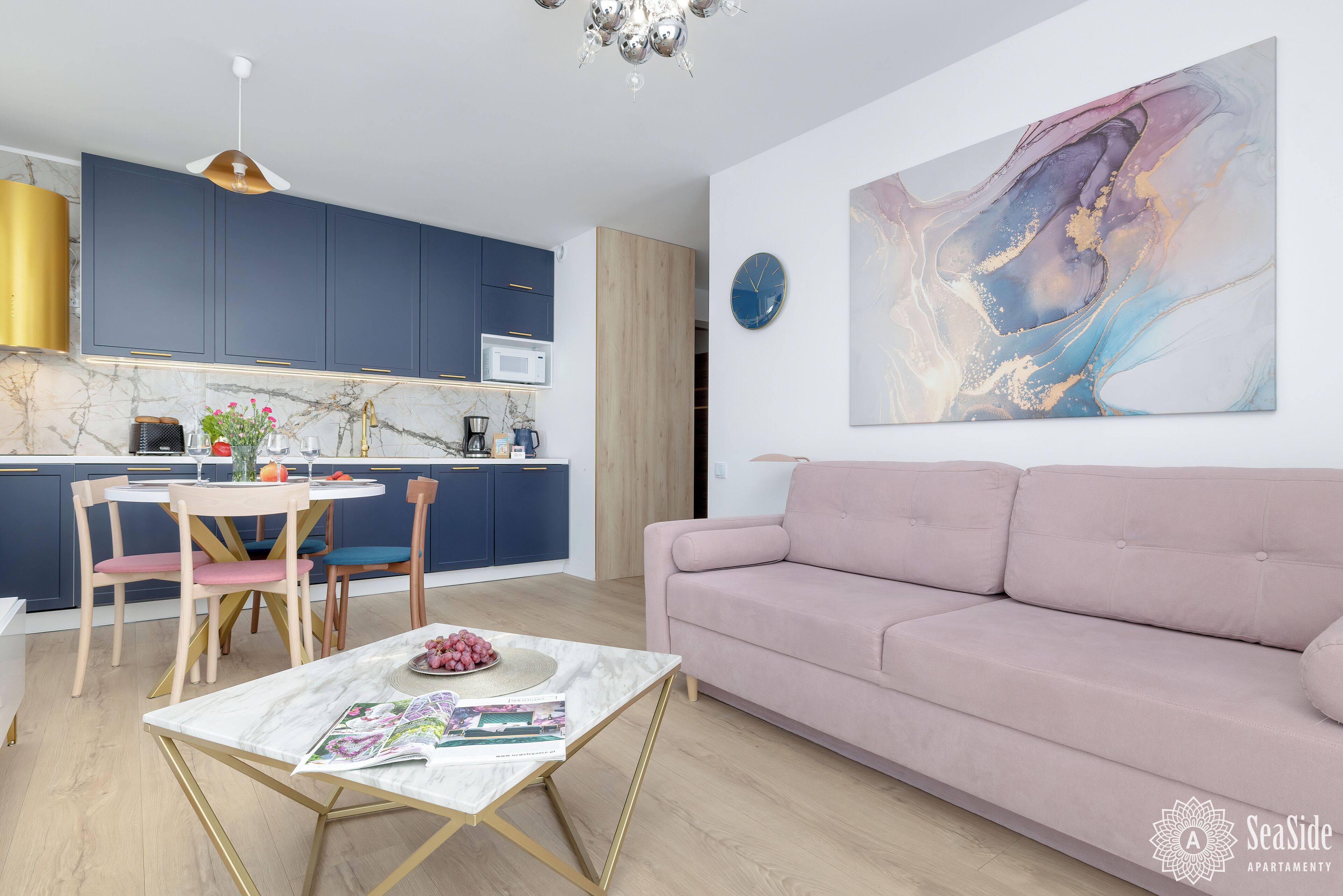 Ein modernes Wohnzimmer mit einem rosafarbenen Sofa, einem runden Tisch mit Zeitschriften und einer Küchenzeile mit blauen Schränken und einer weißen Arbeitsplatte.