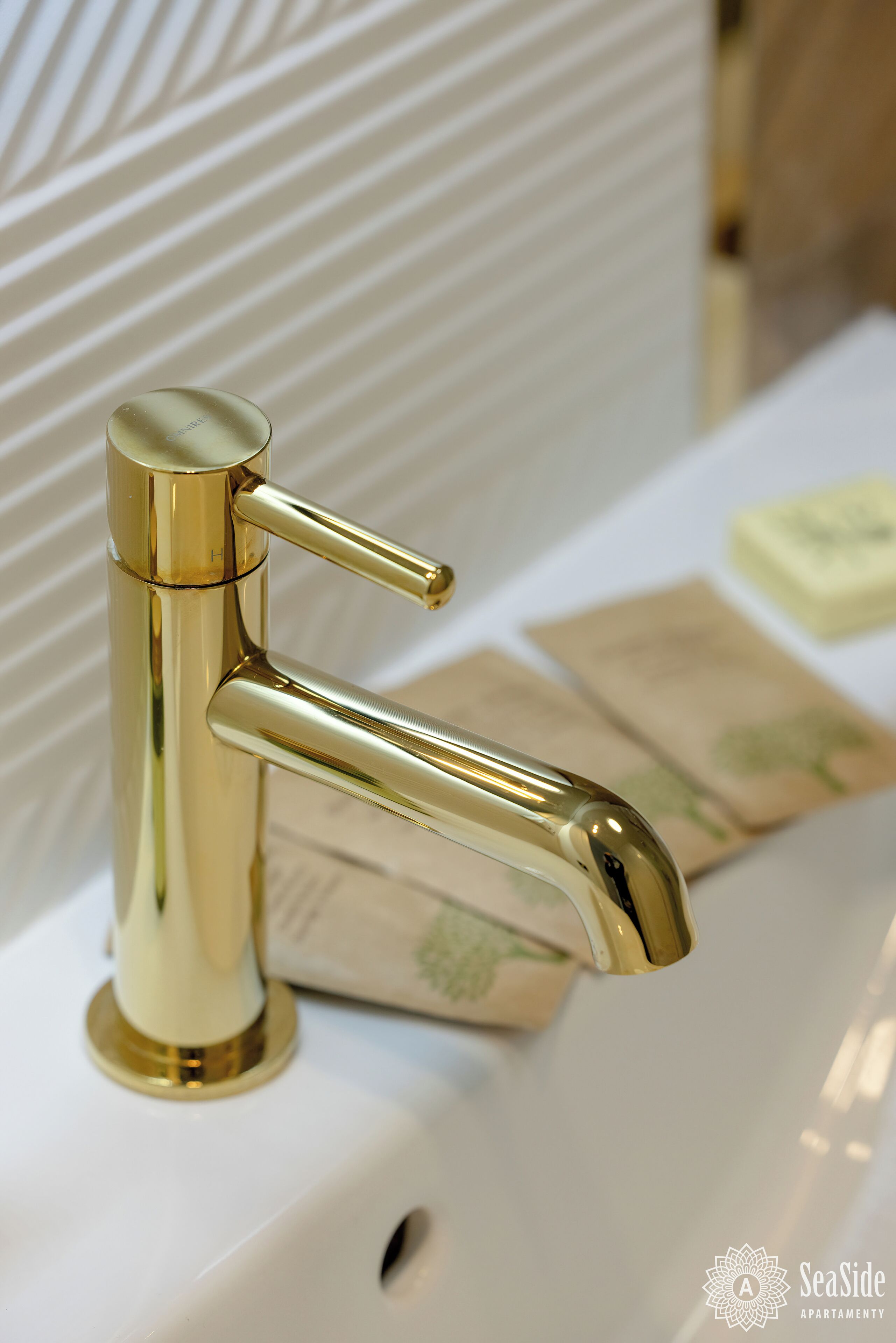 Un grifo dorado sobre un lavabo blanco, con una pared decorada al fondo.