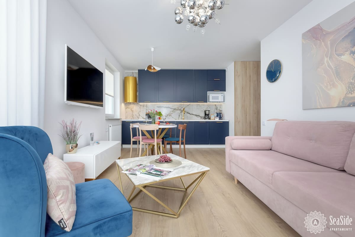 A modern living room with a blue sofa, a pink sofa, and a marble coffee table. There is a kitchen in the background with dark blue cabinets and a white microwave.