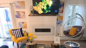 A living room with a blue sofa, yellow armchair, and a hanging chair. There is a flat-screen TV mounted above a fireplace, and artwork on the walls. - Cozy Poconos Spa Retreat Near Hiking, Lakes & Fall Foliage (Tobyhanna)
