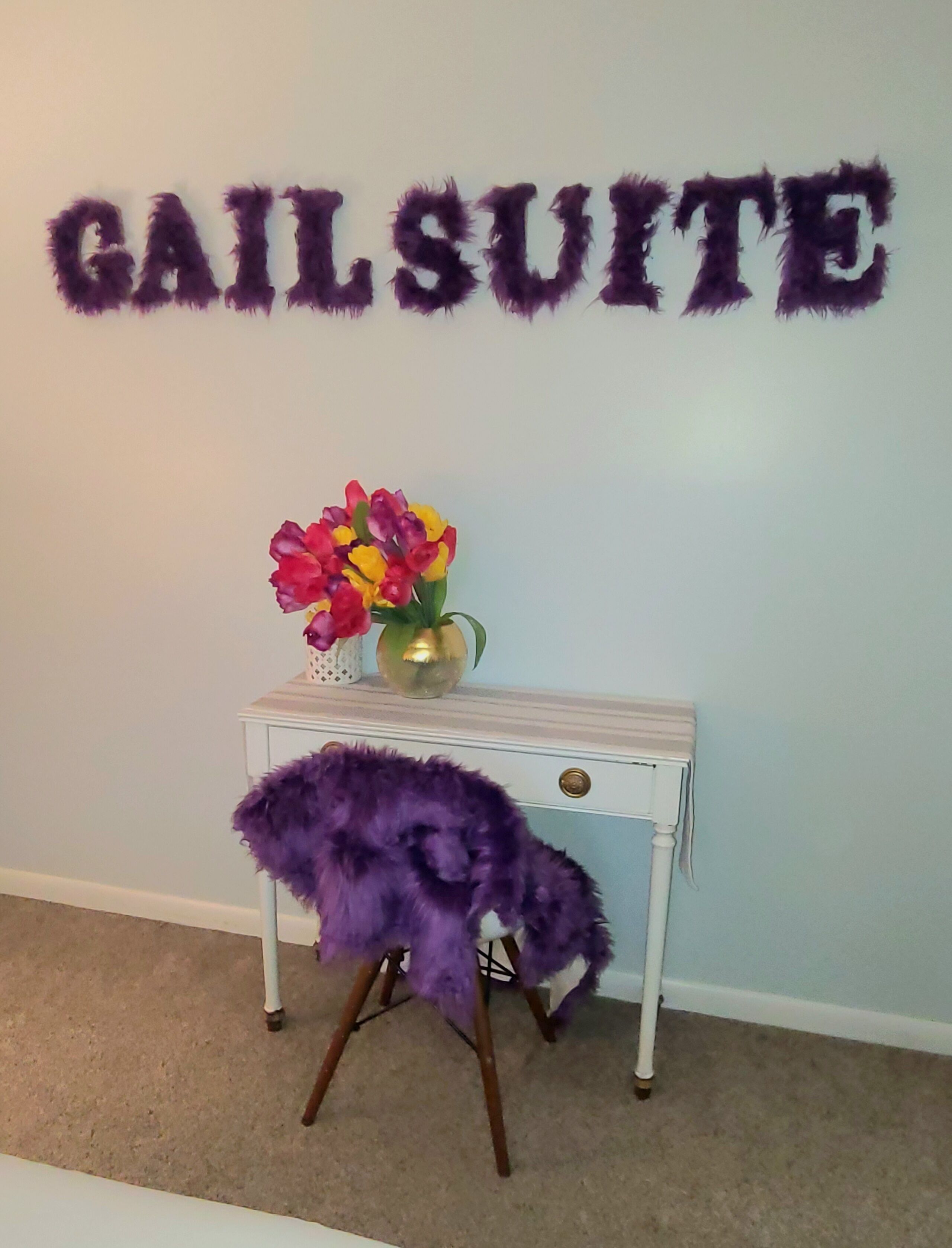 A room with a wall-mounted sign reading "GAIL SUITE" and a small white table with a purple furry chair and a vase of flowers.