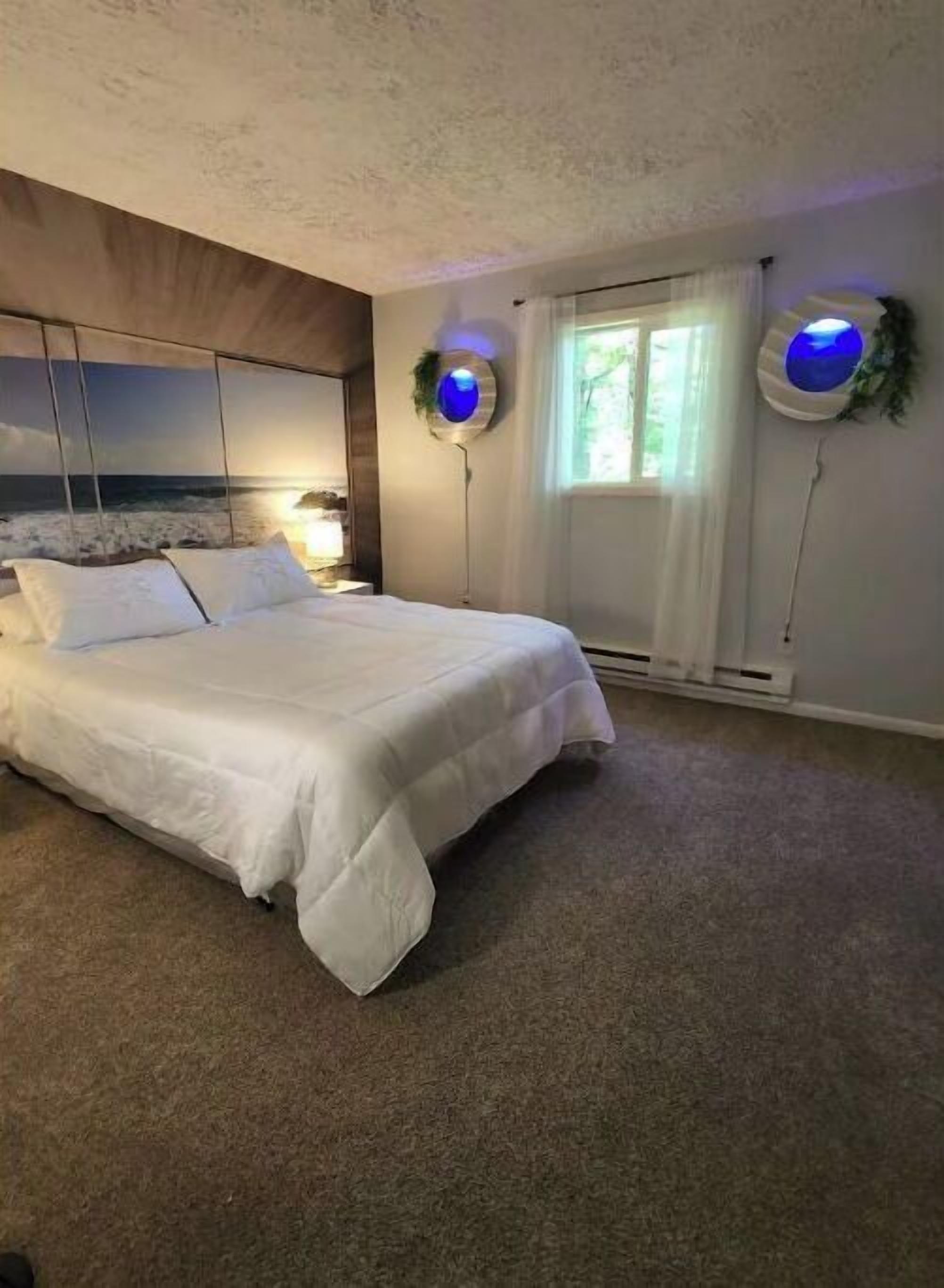 A bedroom with a large bed, a view of the sea through a window, and two wall-mounted lamps with blue lights.