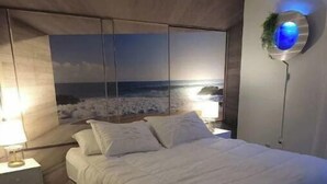 A bedroom with a large bed, a nightstand, a wall-mounted lamp, and a view of the ocean. - Cozy Poconos Spa Retreat Near Hiking, Lakes & Fall Foliage (Tobyhanna)