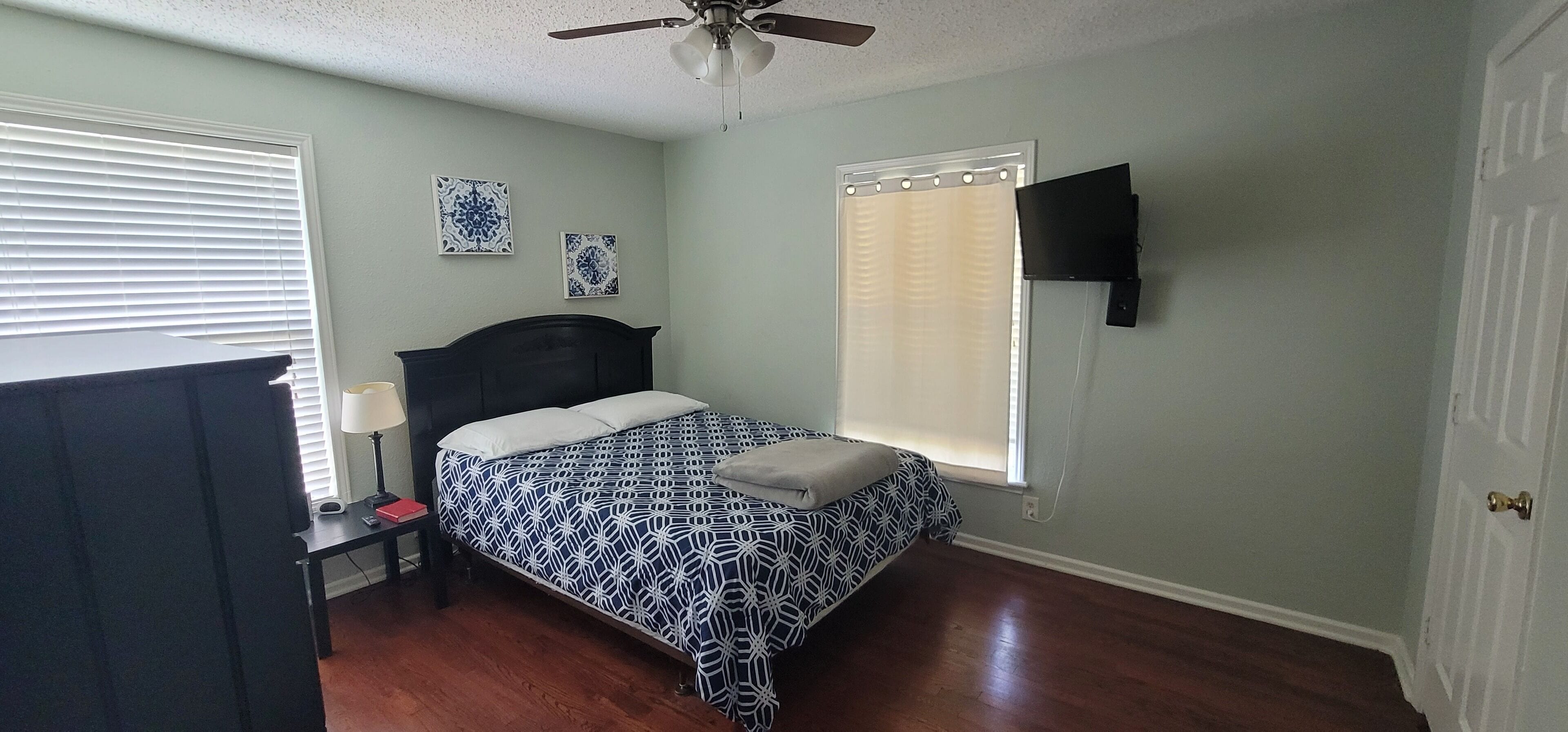 A bedroom with a bed, a nightstand, a lamp, a ceiling fan, and a window with blinds.