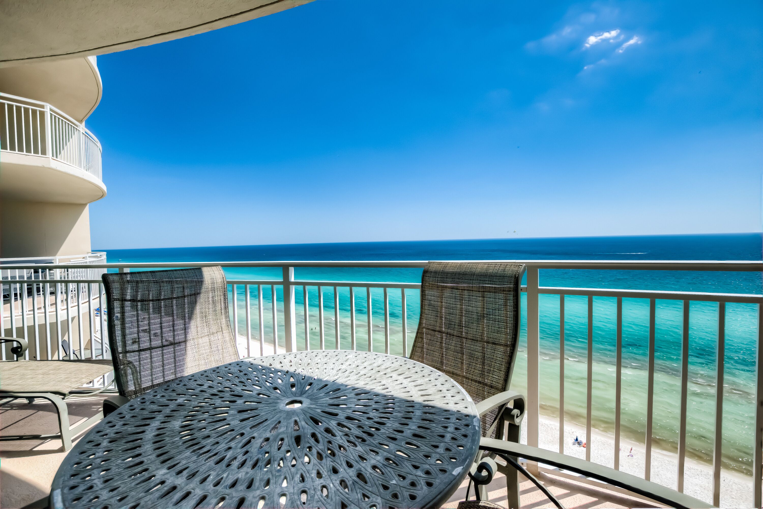 A balcony with a round table and two chairs, overlooking a beach with clear turquoise water and a clear sky.