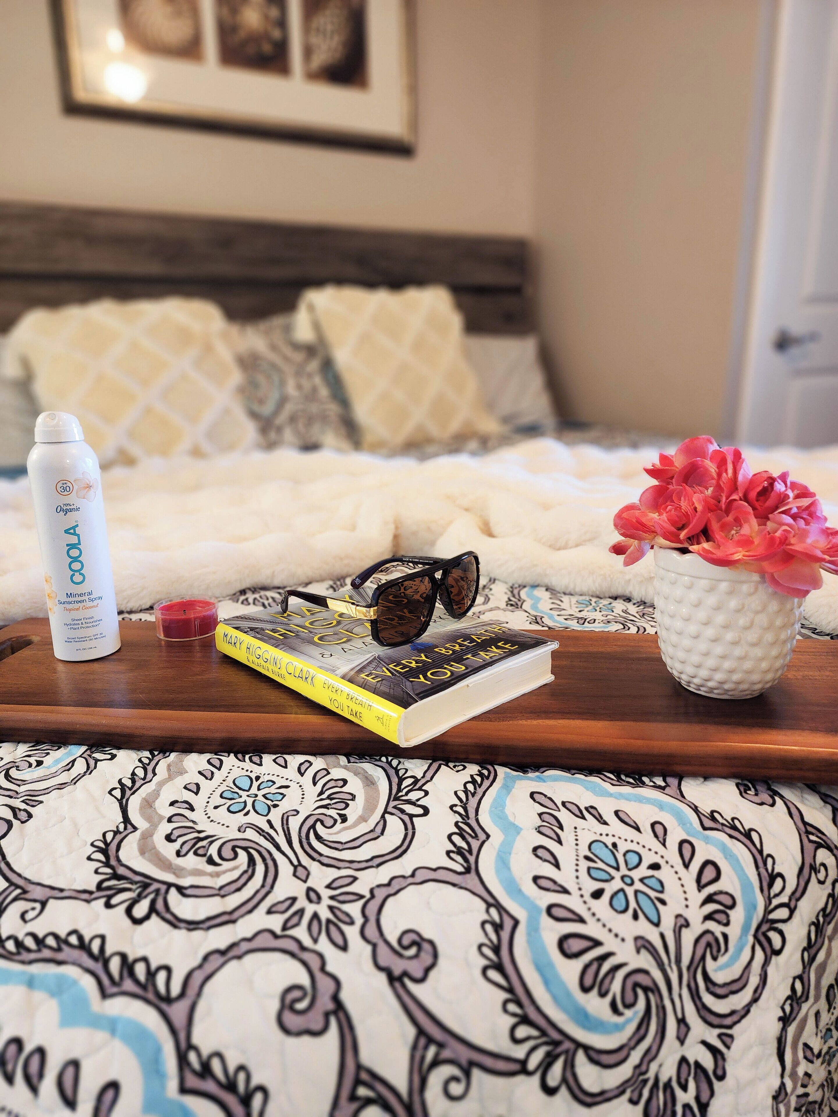 Ein Bett mit einer gemusterten Decke, ein Buch mit dem Titel 'Every Breath You Take' von Mary Higgins Clark, eine Flasche Mineral-Sunscreen-Spray, eine Sonnenbrille und ein kleiner weißer Topf mit rosa Blumen.