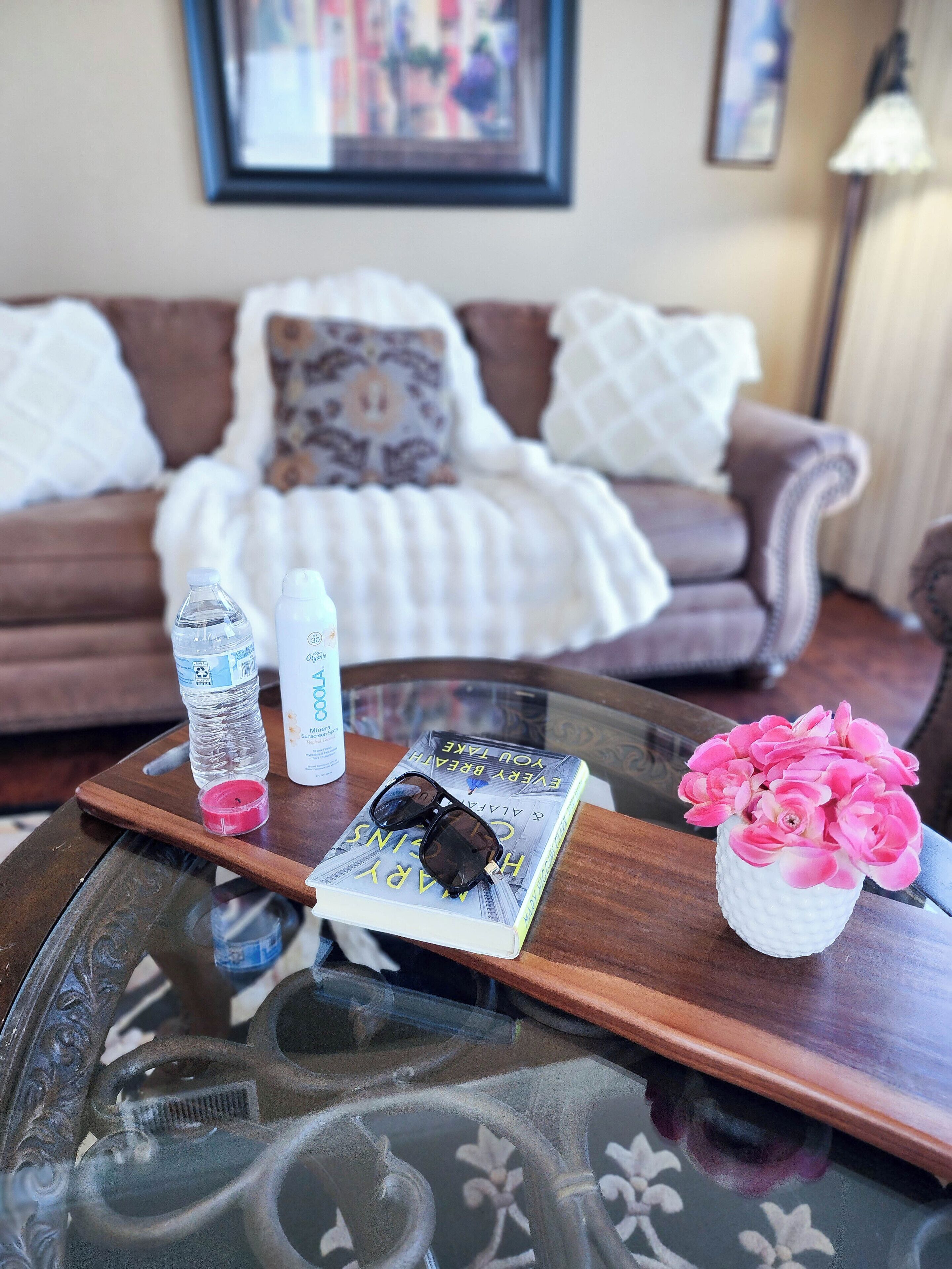 A glass coffee table with a book, sunglasses, a water bottle, a bottle of hand sanitizer, and a vase with pink flowers.
