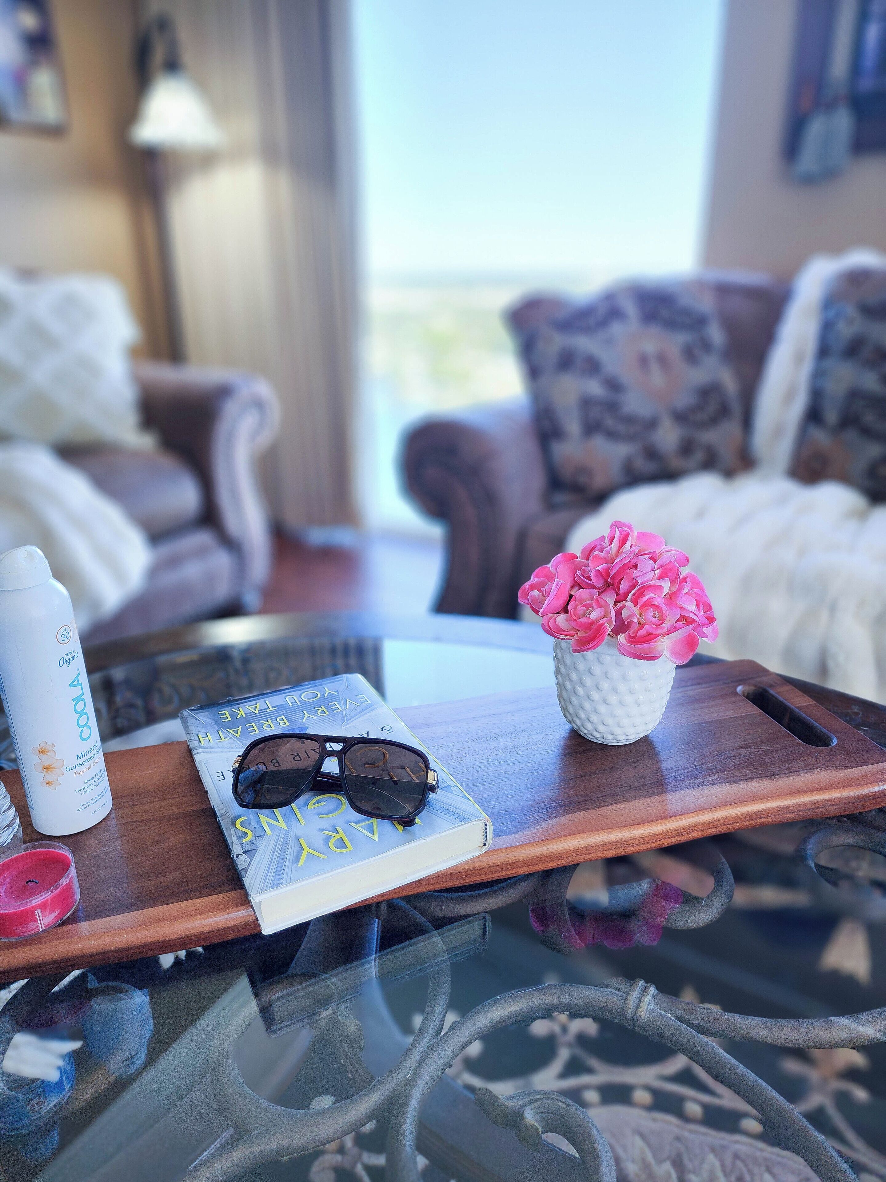 A wooden tray with a book, sunglasses, a water bottle, and a vase with pink flowers on a glass table.