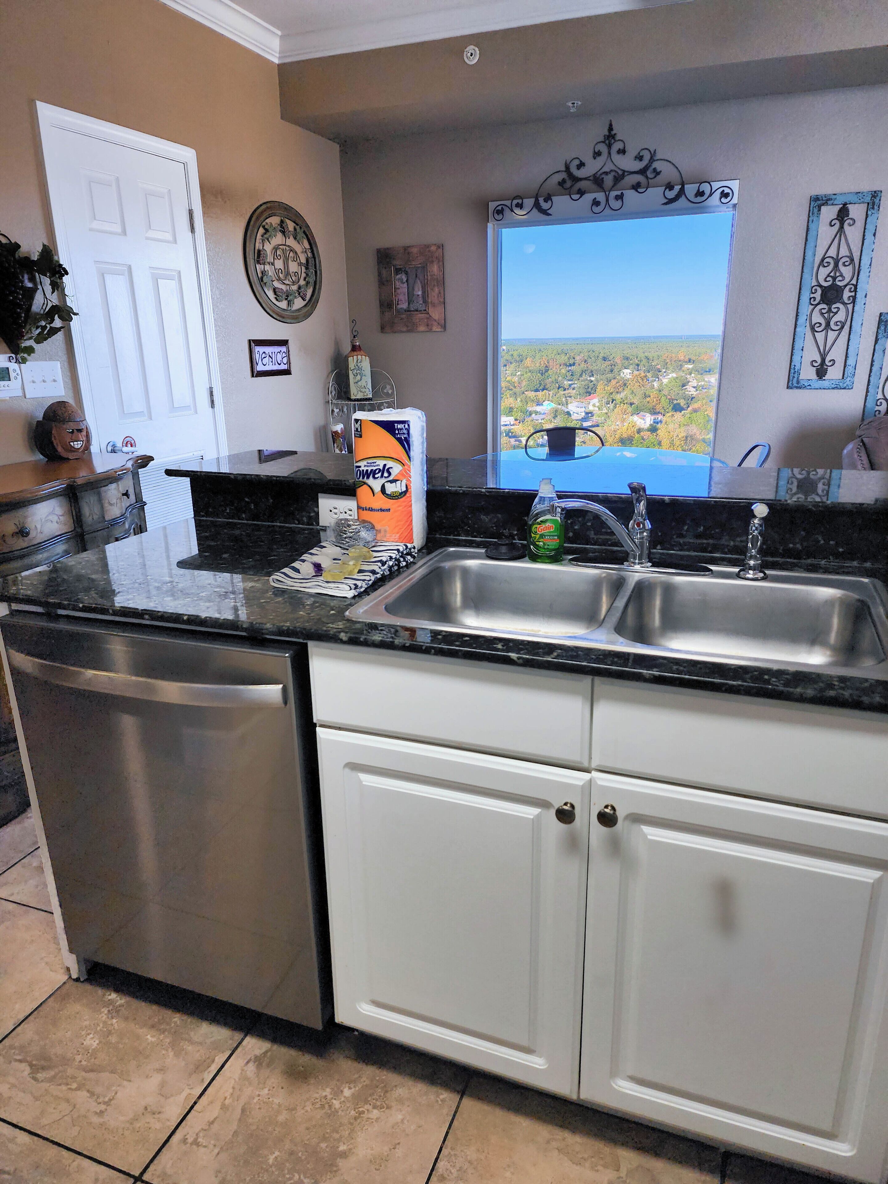 Cocina con lavavajillas, pileta doble y muebles blancos. Se observa una ventana con vista a un pueblo y un objeto decorativo sobre la pileta.