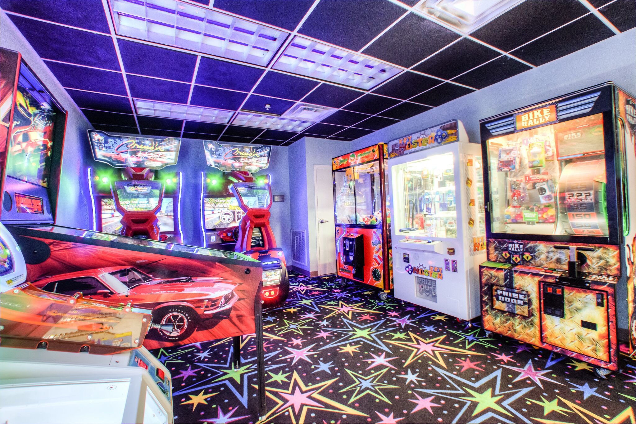 Un salón de juegos muy iluminado con diversas máquinas, incluyendo simuladores de carreras y juegos de premios, y un suelo con diseño de estrellas.