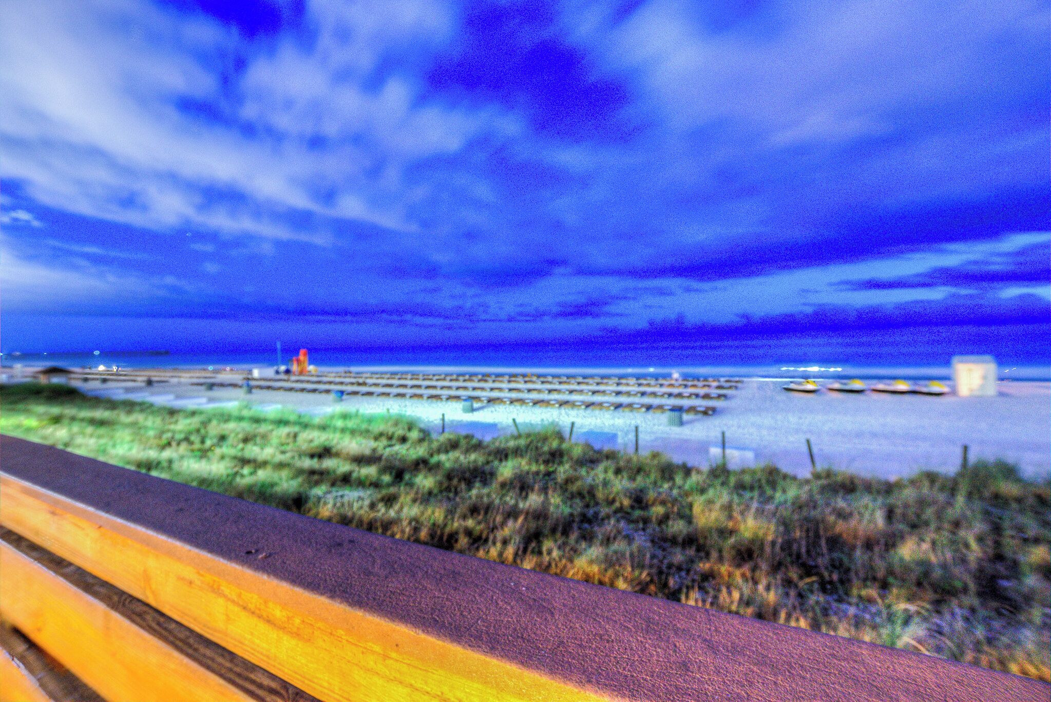 A coastal walkway with a wooden bench, grassy areas, and a pier extending into the water.