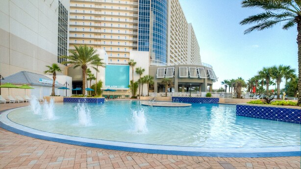 A swimming pool with water fountains, surrounded by palm trees and a brick walkway.