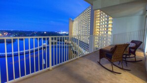 A balcony with a view of a body of water and a cityscape at dusk. There are two wicker rocking chairs and a white railing.