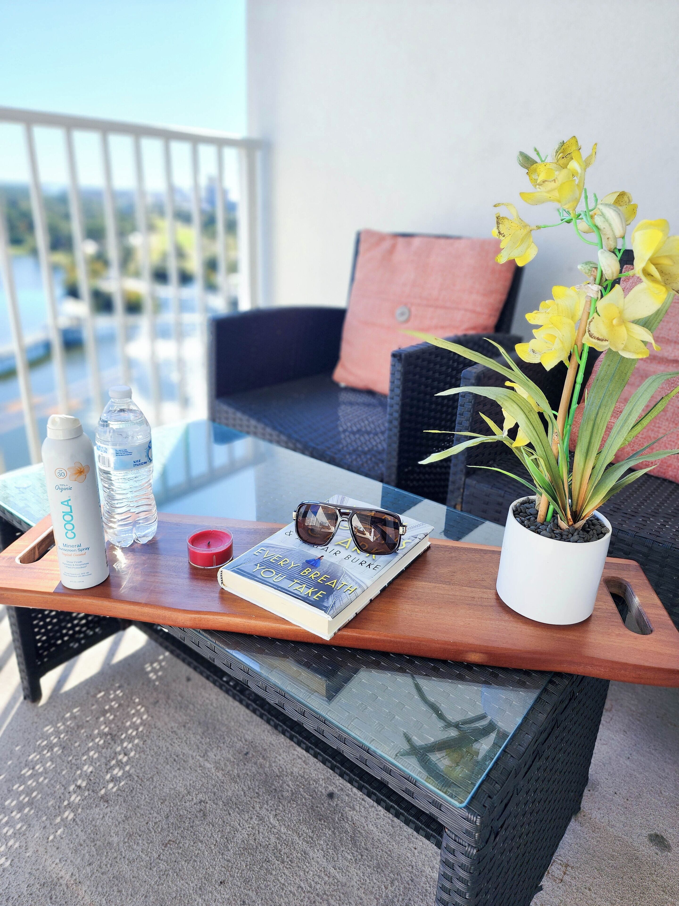 Een balkon met een glazen tafel waarop een waterfles, een boek, een zonnebril en een pot met een plant staan.