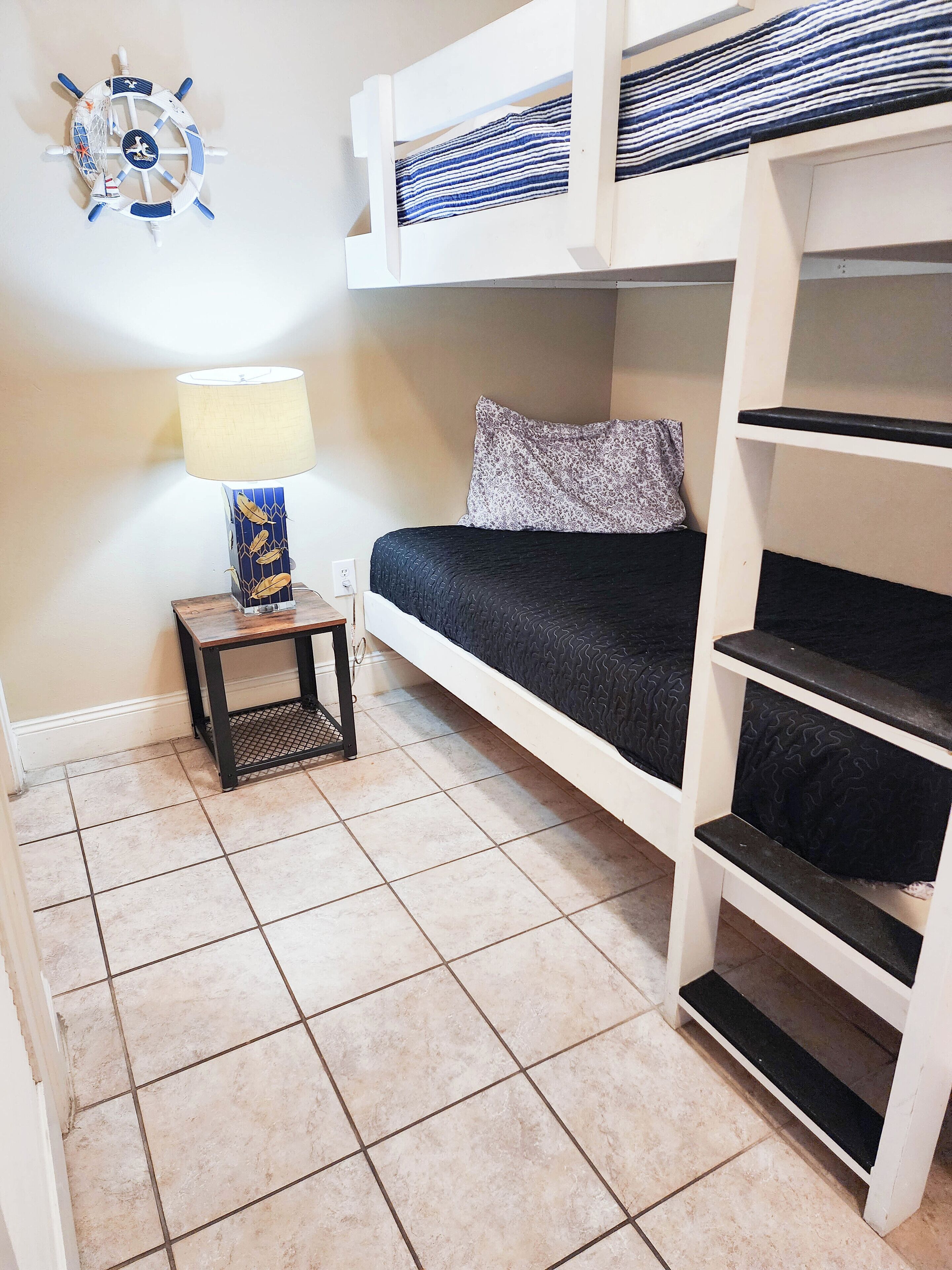 A bunk bed room with a small table and lamp, a decorative wall piece, and tiled flooring.