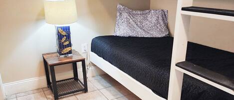 A bunk bed room with a small table and lamp, a decorative wall piece, and tiled flooring.