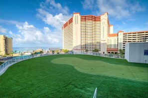 Ein Golfplatz mit gepflegtem Grün, umgeben von einem weißen Zaun, neben einem Hochhaus mit mehreren Etagen und Balkonen.