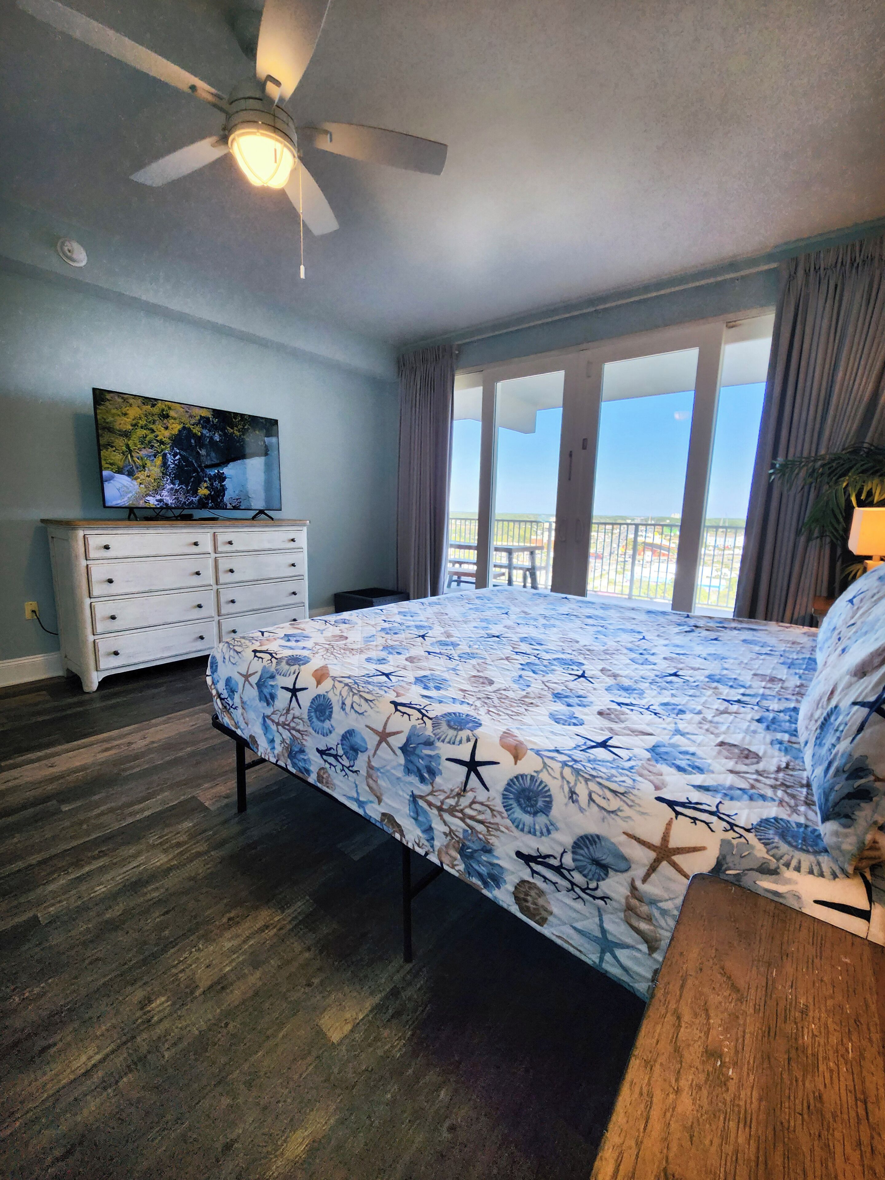 Une chambre avec un lit, un ventilateur de plafond, une télévision, une commode et un balcon avec vue.