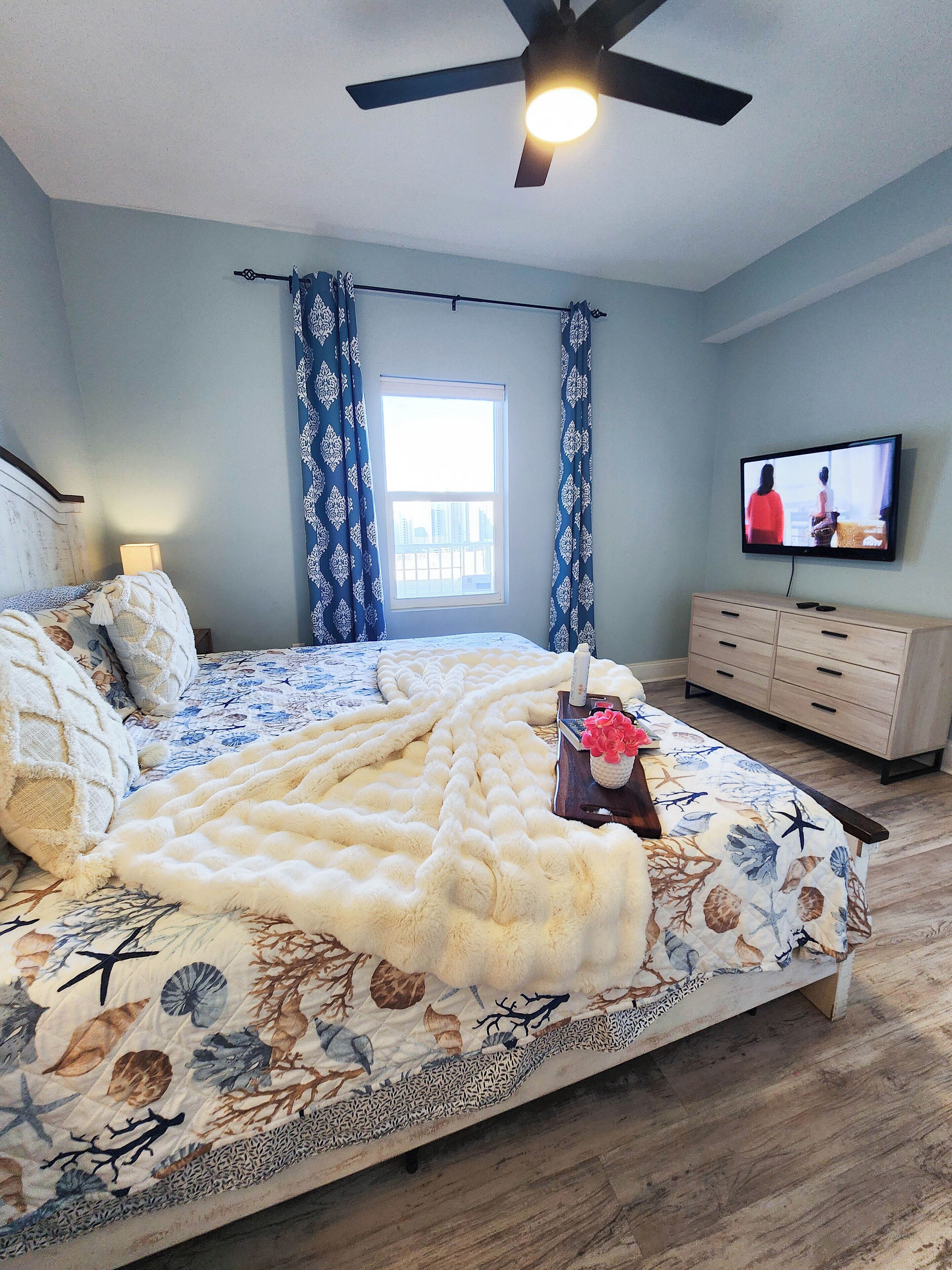 A bedroom with a bed, a ceiling fan, a TV, a dresser, and a window with curtains.