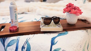 Una cama con colcha blanca y azul estampada con motivos florales, una bandeja de madera con una botella de protector solar, gafas de sol y un pequeño jarrón con flores rosadas.