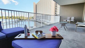 A balcony with blue cushioned seating, a small table with a book, sunglasses, and a water bottle. A potted plant with pink flowers is also visible.