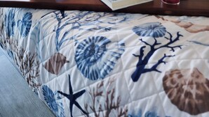 A neatly made bed with a floral-patterned duvet, a bedside table with a book, a pair of sunglasses, a candle, and a water bottle.