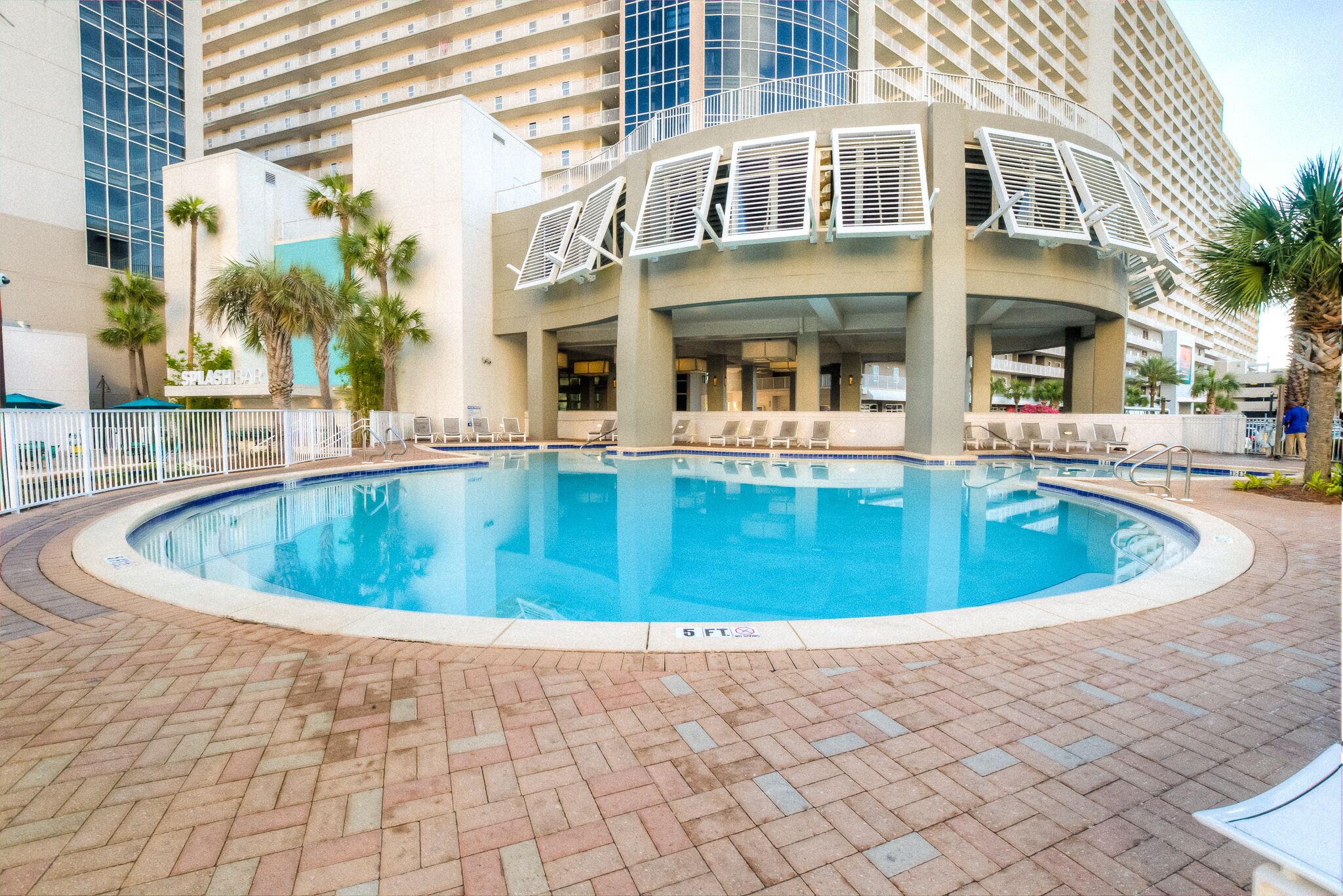 A swimming pool with lounge chairs and palm trees, surrounded by a brick walkway and a building with a modern design.