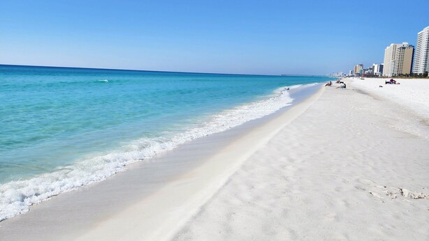 Een breed zandstrand met helderblauw water en enkele gebouwen op de achtergrond.
