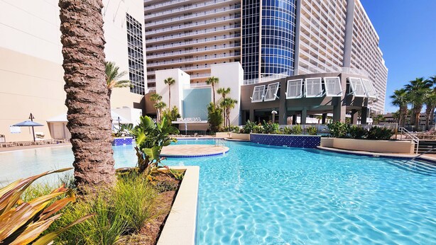 A hotel pool area with a clear blue pool, palm trees, and a modern building with balconies in the background. - Perfect views! Incredible experience! Best onsite restaurants and shops! (Panama City Beach)