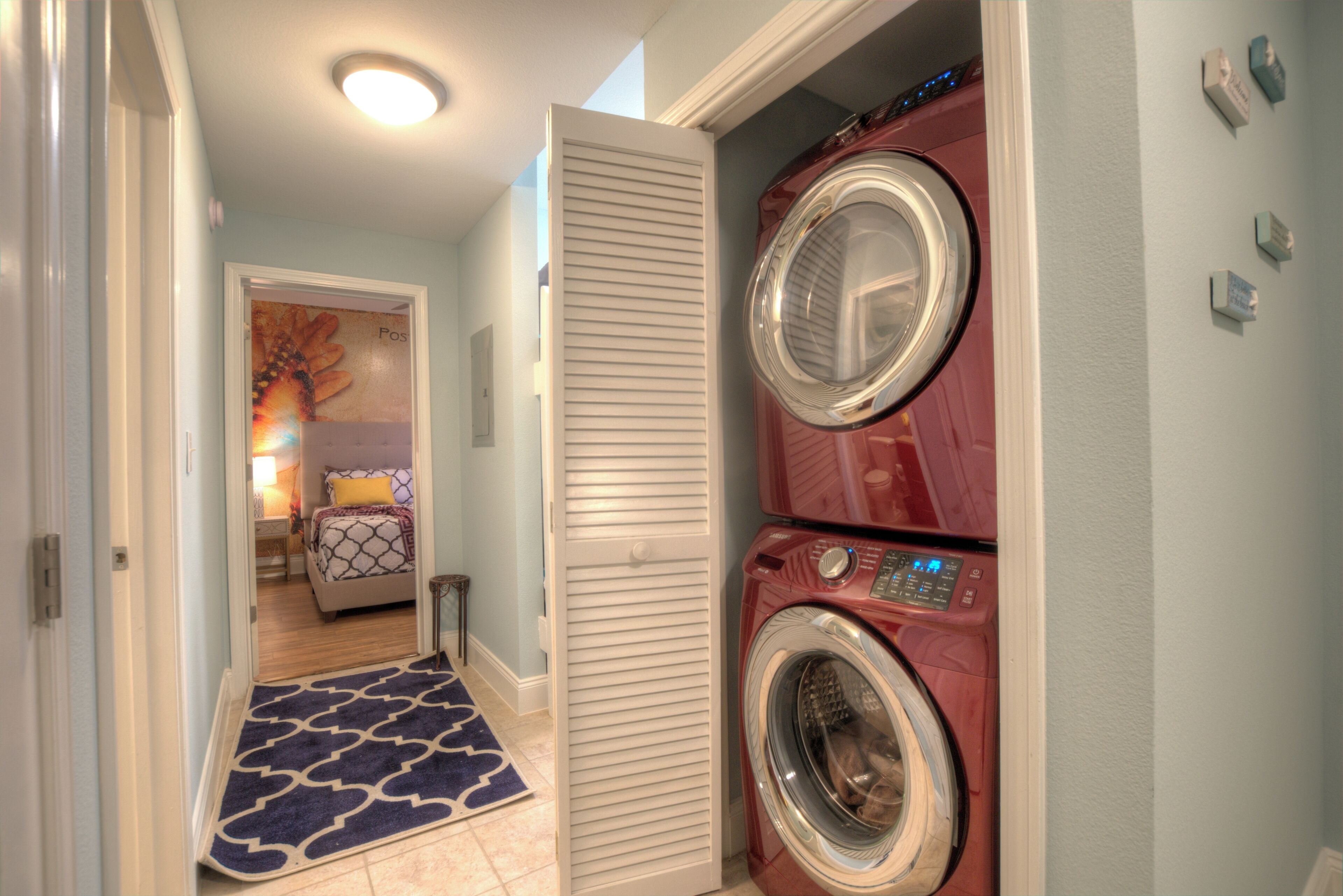 A laundry room with two stacked front-loading washers and dryers, a blue patterned rug, and a door leading to a bedroom with a bed and a nightstand.