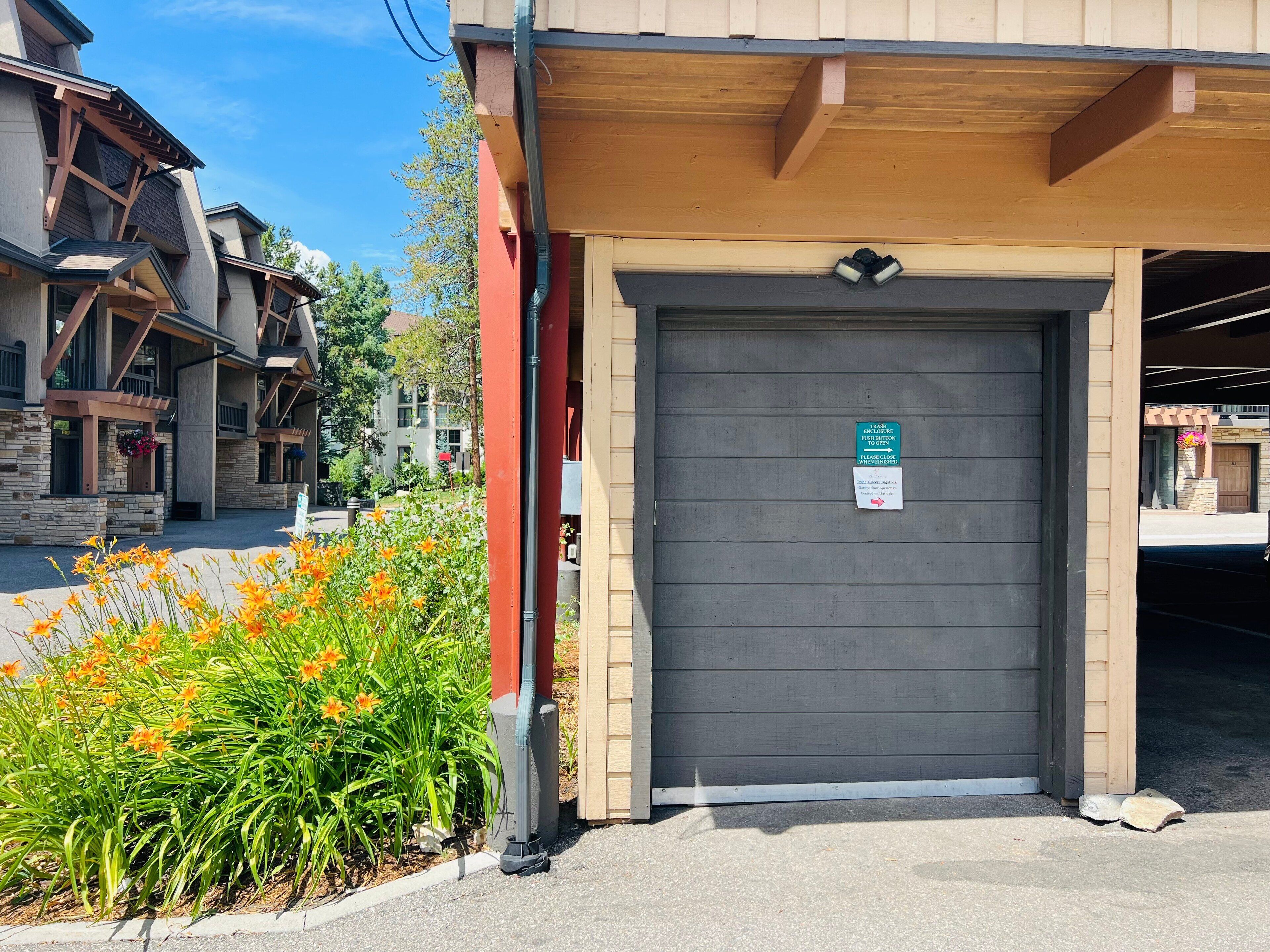 Entrada de um edifício com uma porta de garagem fechada, um contentor de lixo e uma placa a indicar uma área para depositar o lixo.