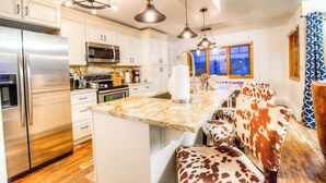 A modern kitchen with white cabinets, stainless steel appliances, and a marble countertop. There are pendant lights, a large window, and a seating area with cowhide chairs.