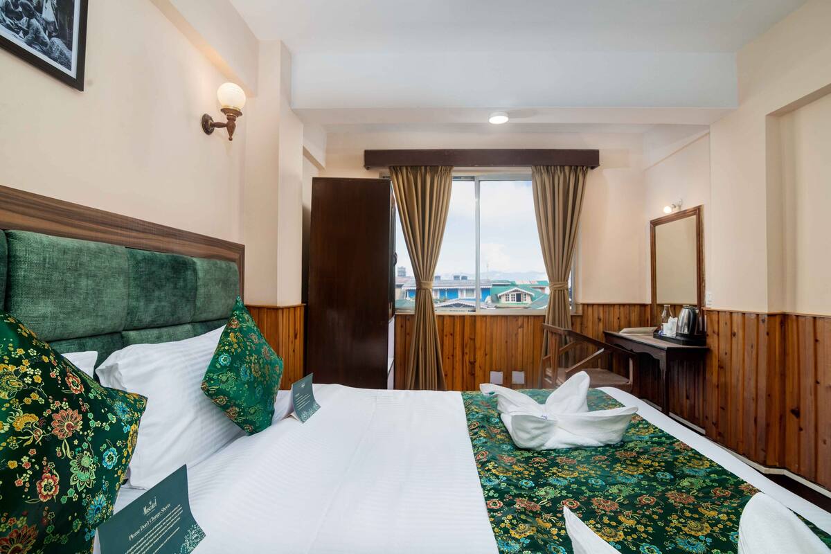 A hotel room with a large bed, a wooden headboard, a view of buildings through the window, a desk with a mirror, and a wooden wall paneling.