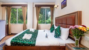 A hotel room with a large bed, a wooden headboard, a nightstand with a vase of flowers, and a window with curtains. - Muscatel Roma (Darjeeling)