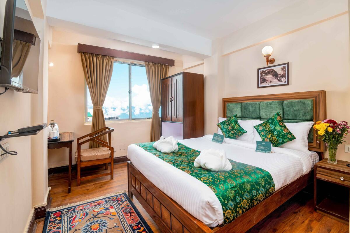 A hotel room with a large bed, a wooden headboard, a desk, a chair, a window with curtains, a nightstand, and a vase with flowers.