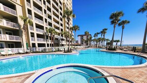 A large outdoor swimming pool with a surrounding brick deck, palm trees, and a multi-story building with balconies. - Bring your Pet! Amazing 2nd floor unit! Outstanding Views and Amenities! (Panama City Beach)