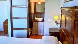 A bedroom with a wooden door, a bed with a white bedspread, a bedside table, and a nightstand. - Belvedere Farmhouse: 2 Bedrooms, 2 Bathrooms, AC | Bolgheri & Sea at 10 minutes (Castagneto Carducci)