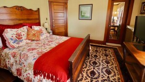 A bedroom with a wooden bed, a red blanket, a patterned rug, a wooden dresser, and a door leading to another room. - The Marshall Room w/ queen bed (Niagara Falls)