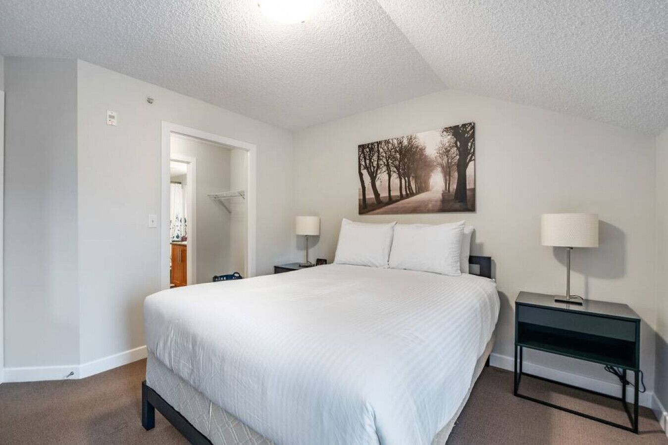 A bedroom with a large bed, two bedside tables with lamps, a door leading to a bathroom, and a painting on the wall.