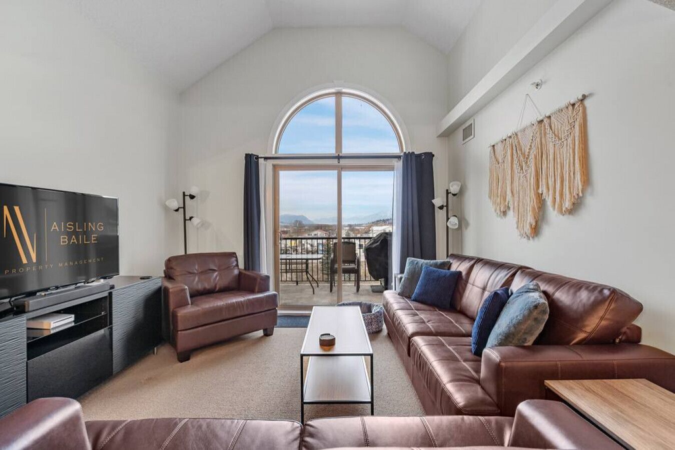 A modern living room with a leather sofa, a coffee table, and a flat-screen TV. There is a balcony with a view of the city.