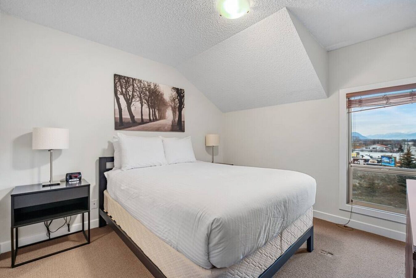 Une chambre avec un grand lit, une table de chevet, une lampe, un tableau au mur et une fenêtre donnant sur un beau paysage.